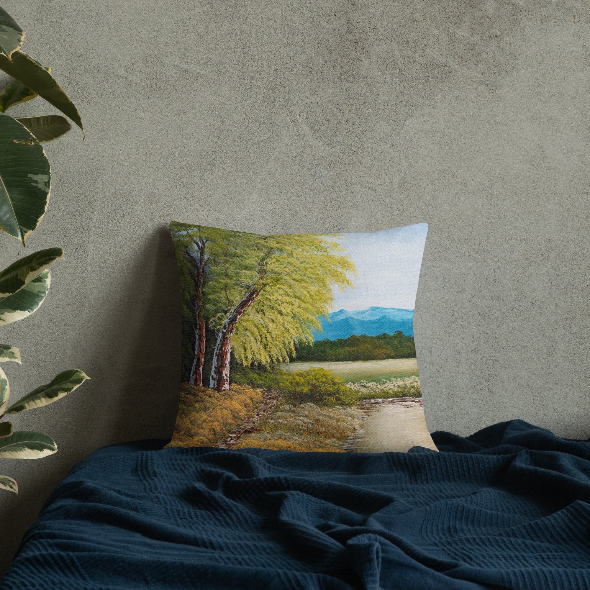 Exclusive and decorative premium designer cushion with an all-over print of a painted landscape with a large glade and a big green tree under a blue sky, placed on a blue blanket.