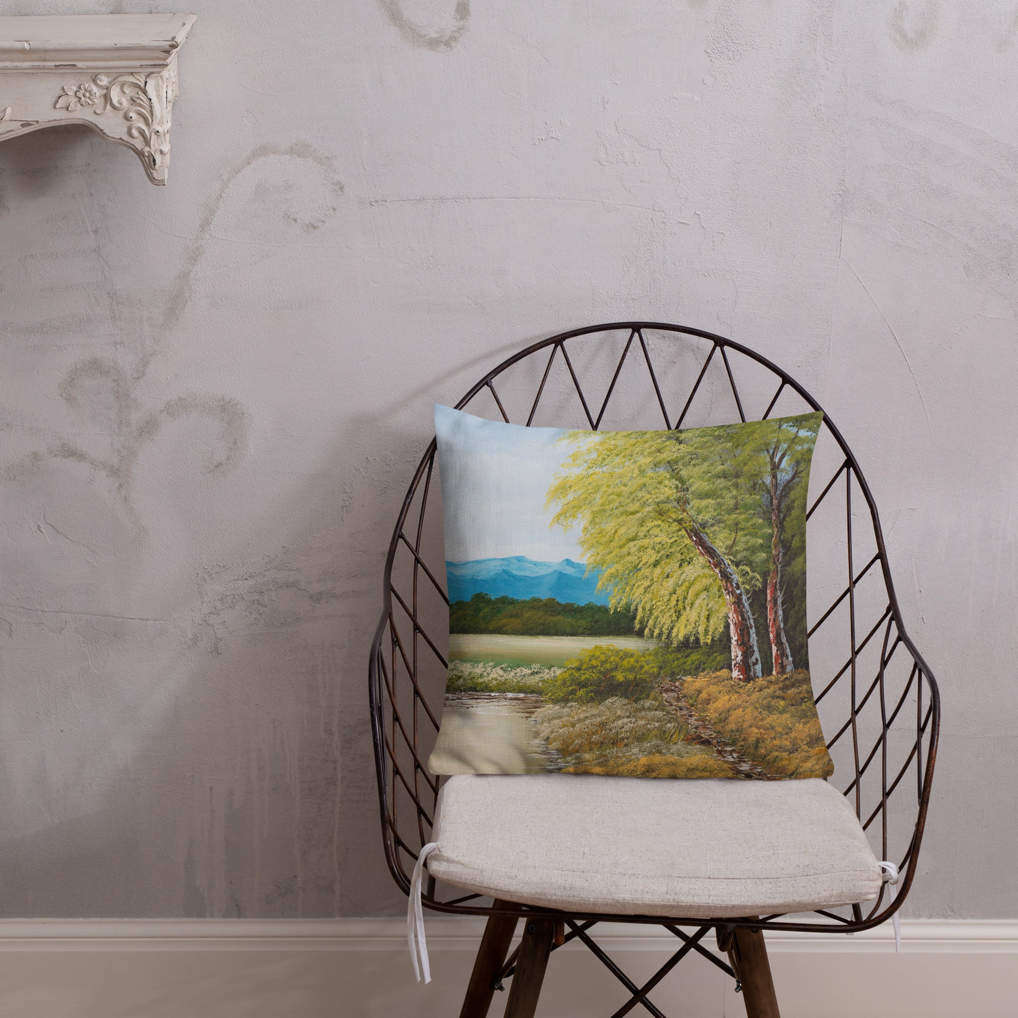 Exclusive and decorative premium designer cushion with an all-over print of a painted landscape with a large glade and a big green tree under a blue sky, placed on a chair.