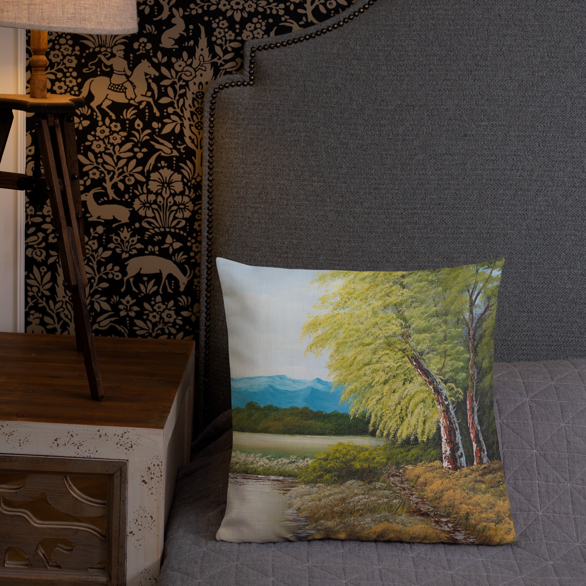Exclusive and decorative premium designer cushion with an all-over print of a painted landscape with a large glade and a big green tree under a blue sky, placed on a bed.