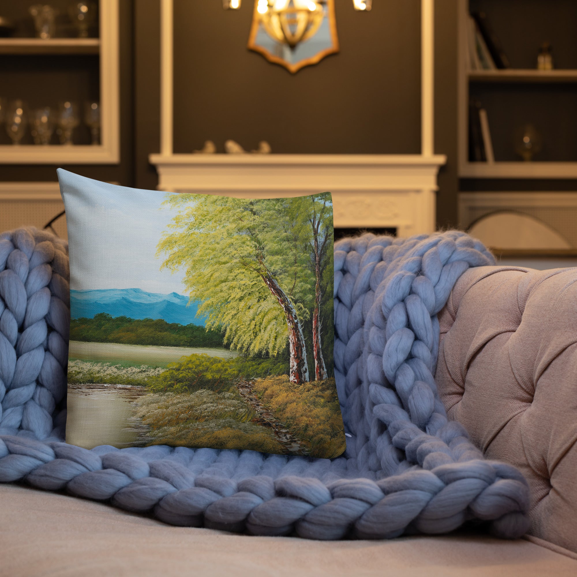 Exclusive and decorative premium designer cushion with an all-over print of a painted landscape with a large glade and a big green tree under a blue sky, placed on a sofa.