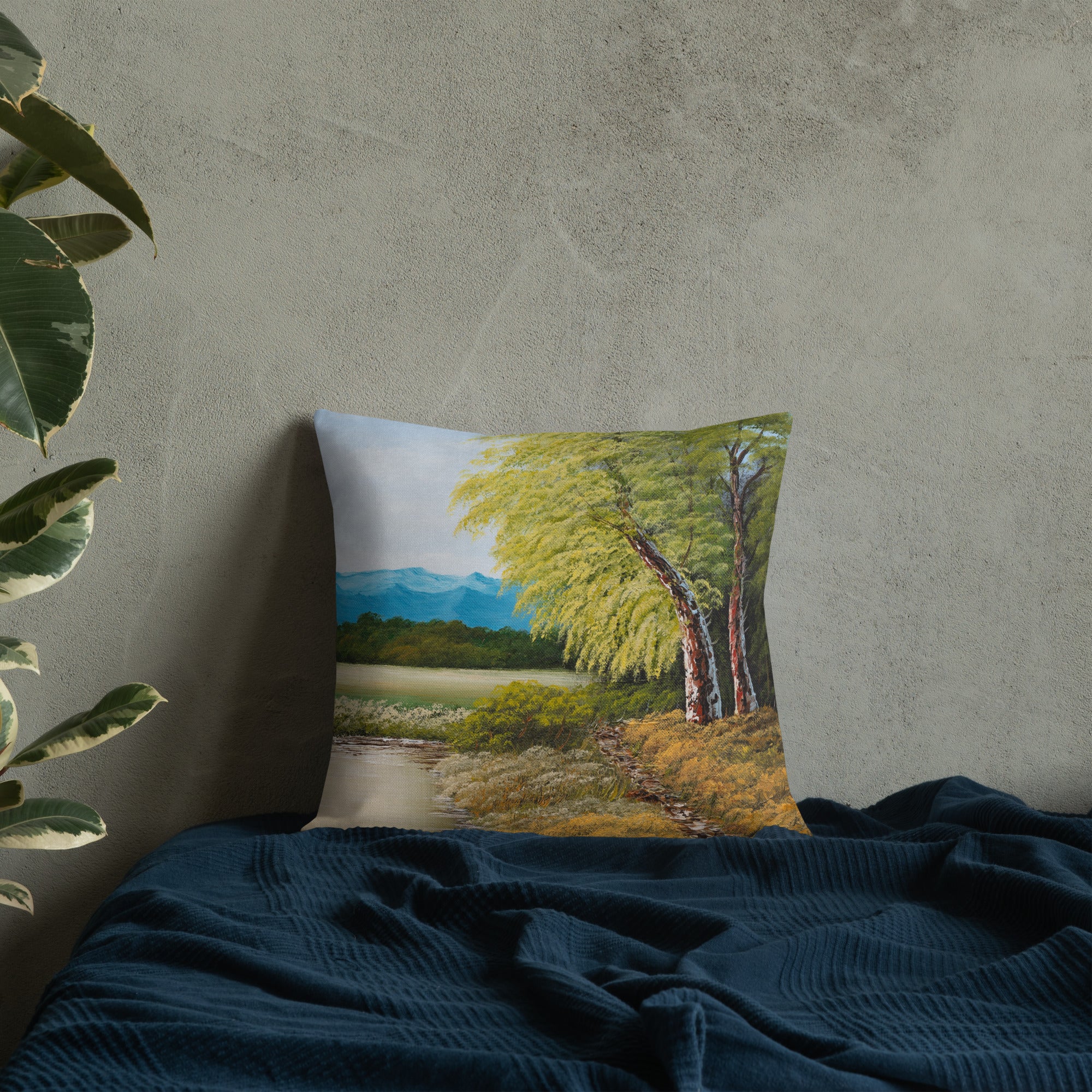 Exclusive and decorative premium designer cushion with an all-over print of a painted landscape with a large glade and a big green tree under a blue sky, placed on a bed.