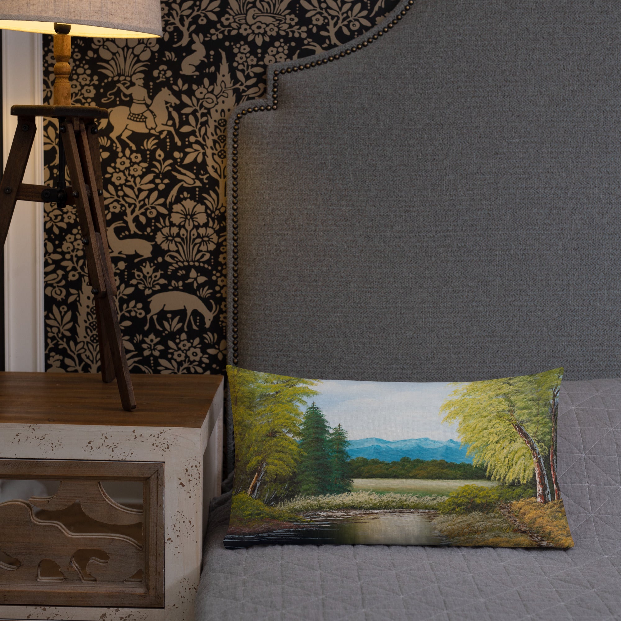 Exclusive and decorative premium designer cushion with an all-over print of a painted landscape with a large glade and a big green tree under a blue sky, placed on a bed.