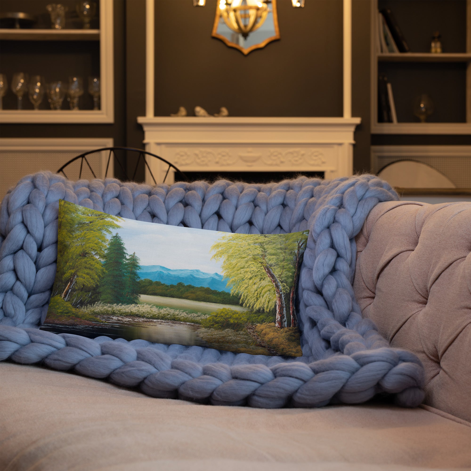 Exclusive and decorative premium designer cushion with an all-over print of a painted landscape with a large glade and a big green tree under a blue sky, placed on a sofa.