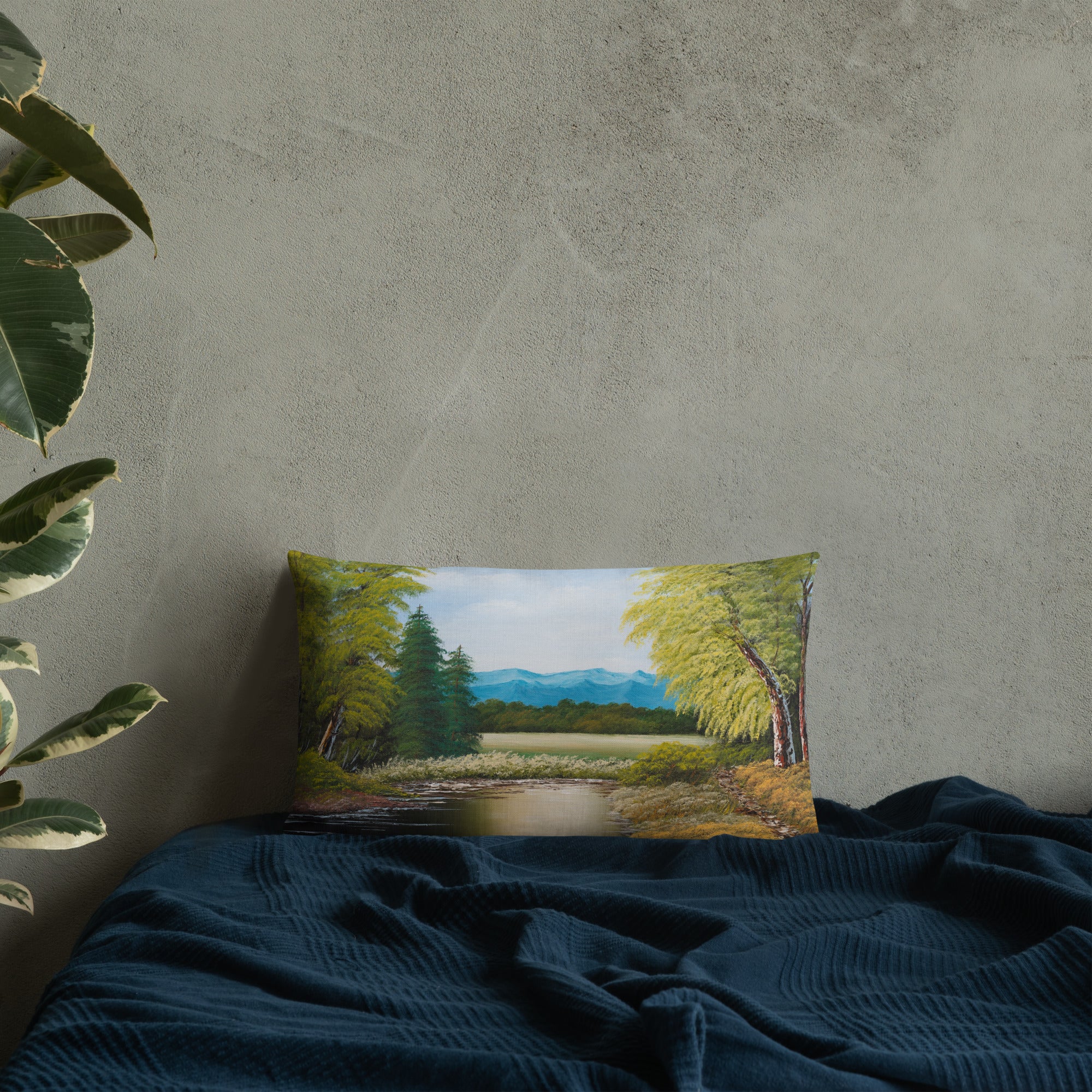 Exclusive and decorative premium designer cushion with an all-over print of a painted landscape with a large glade and a big green tree under a blue sky, placed on a bed.