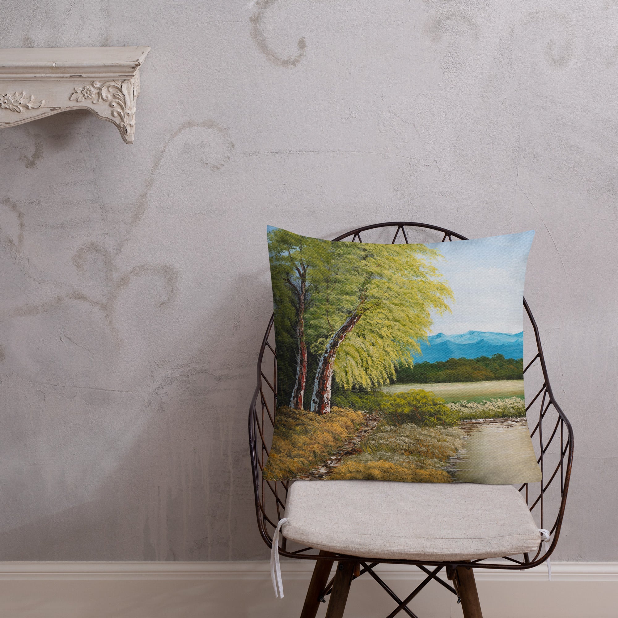 Exclusive and decorative premium designer cushion with an all-over print of a painted landscape with a large glade and a big green tree under a blue sky, placed on a chair.
