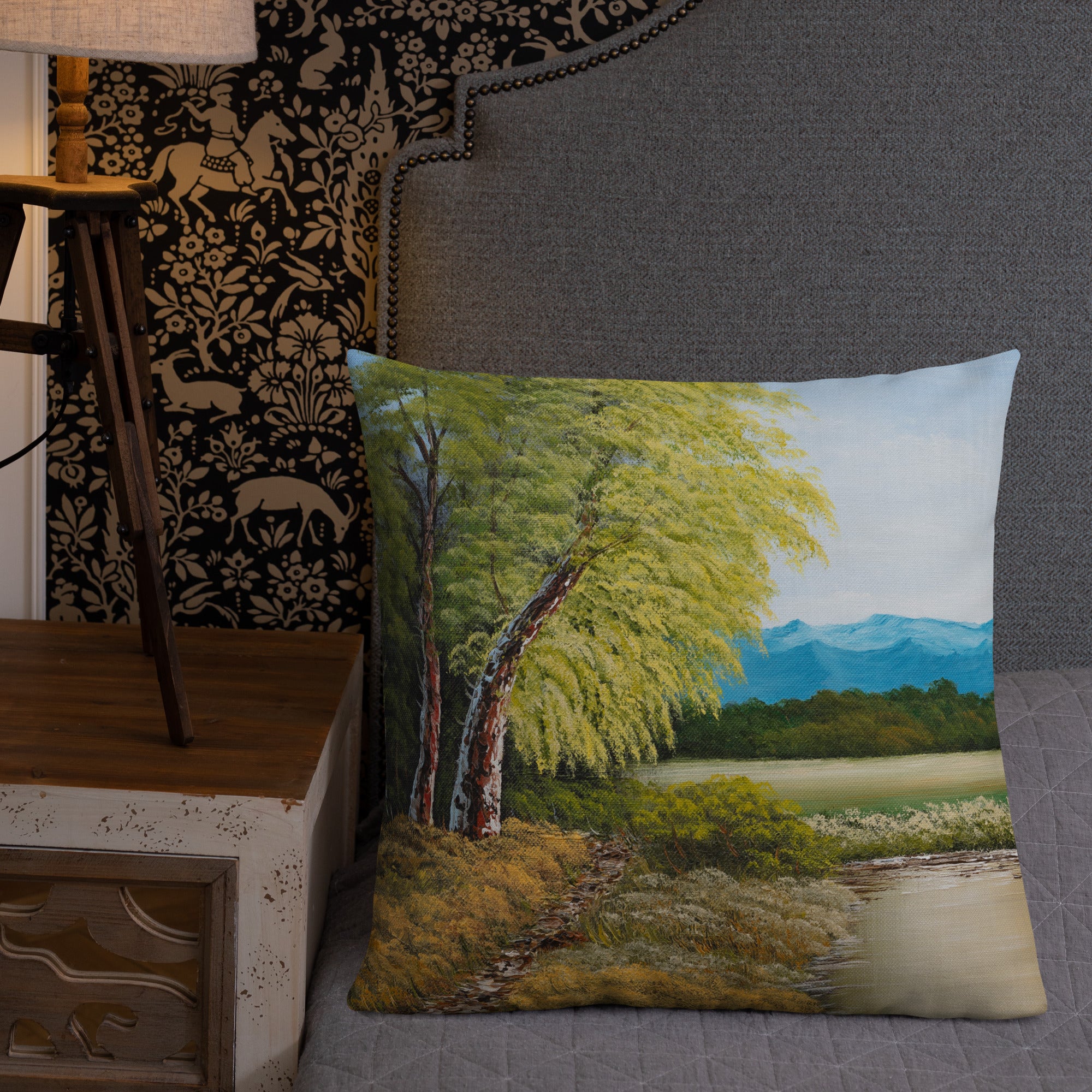 Exclusive and decorative premium designer cushion with an all-over print of a painted landscape with a large glade and a big green tree under a blue sky, placed on a bed.