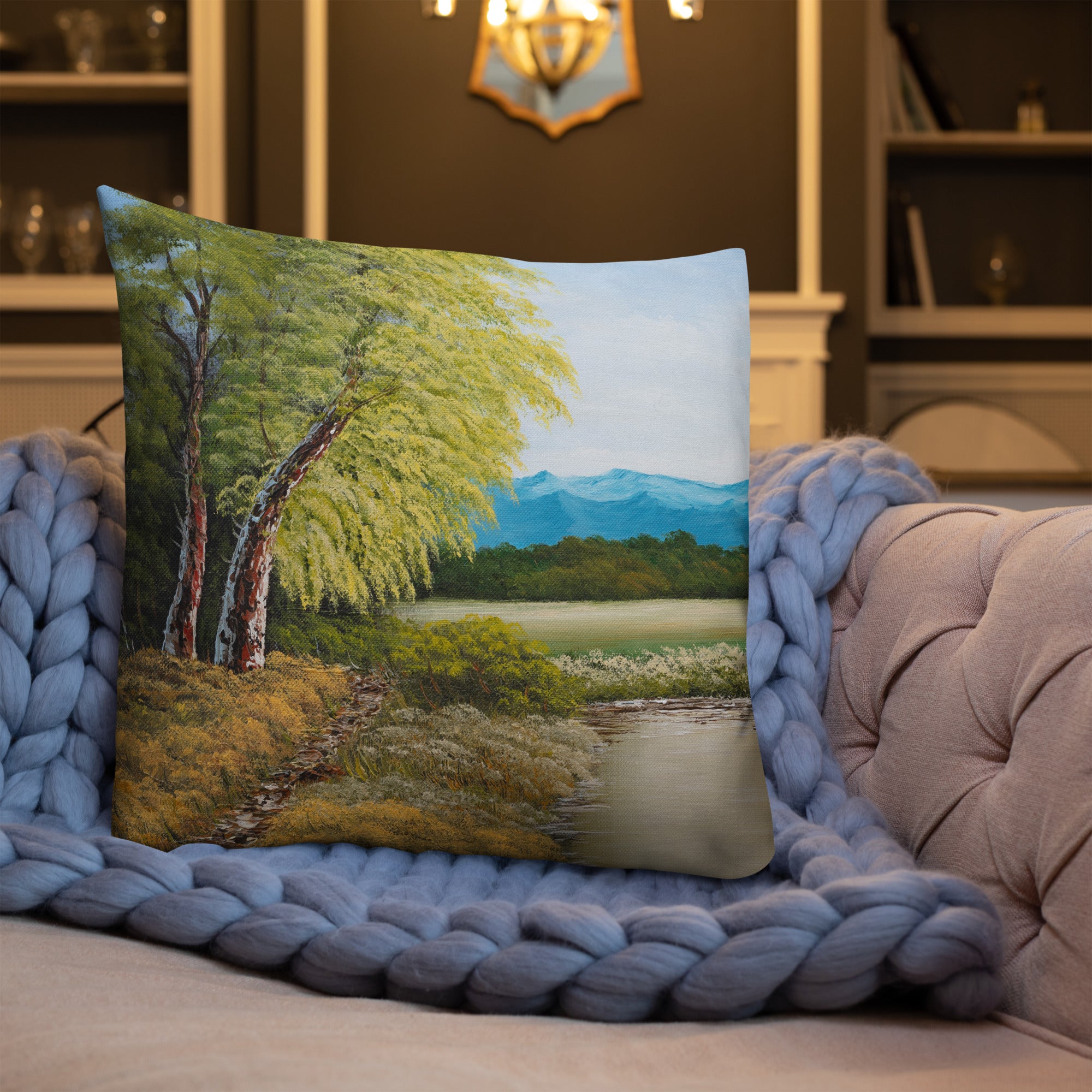 Exclusive and decorative premium designer cushion with an all-over print of a painted landscape with a large glade and a big green tree under a blue sky, placed on a sofa.