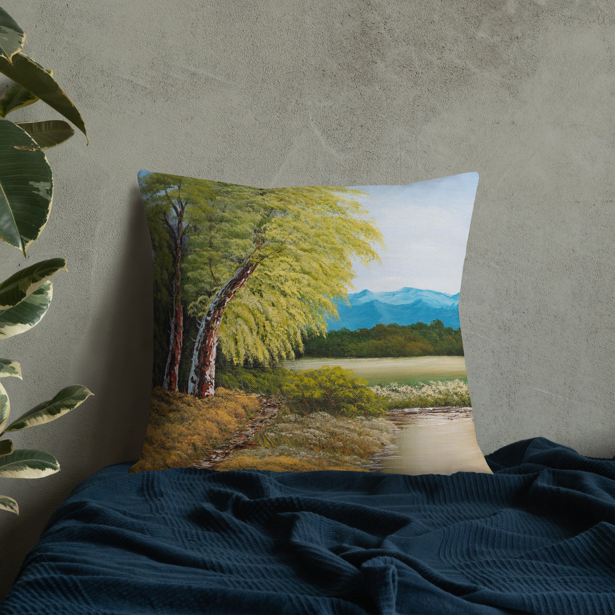 Exclusive and decorative premium designer cushion with an all-over print of a painted landscape with a large glade and a big green tree under a blue sky, placed on a bed.