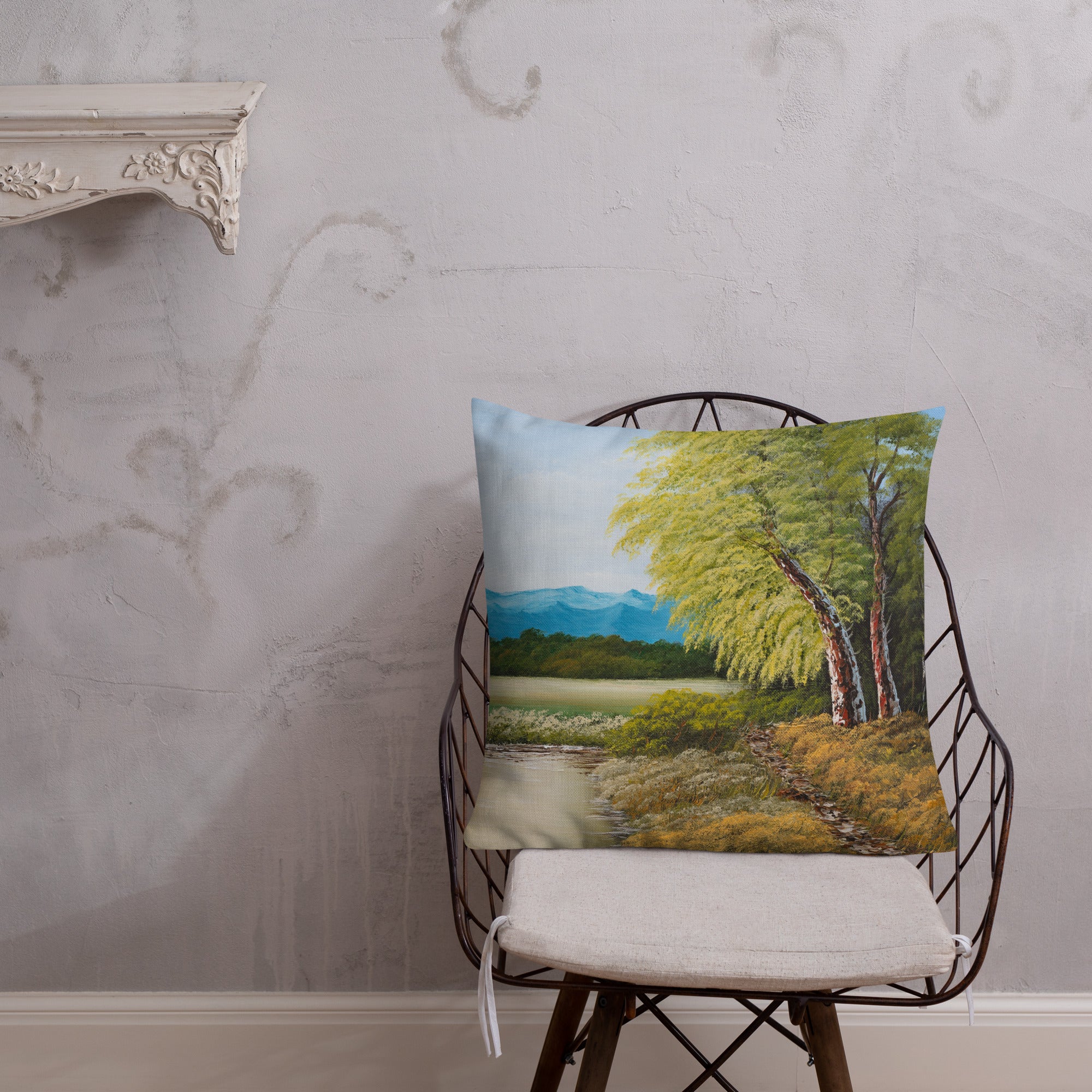Exclusive and decorative premium designer cushion with an all-over print of a painted landscape with a large glade and a big green tree under a blue sky, placed on a chair.