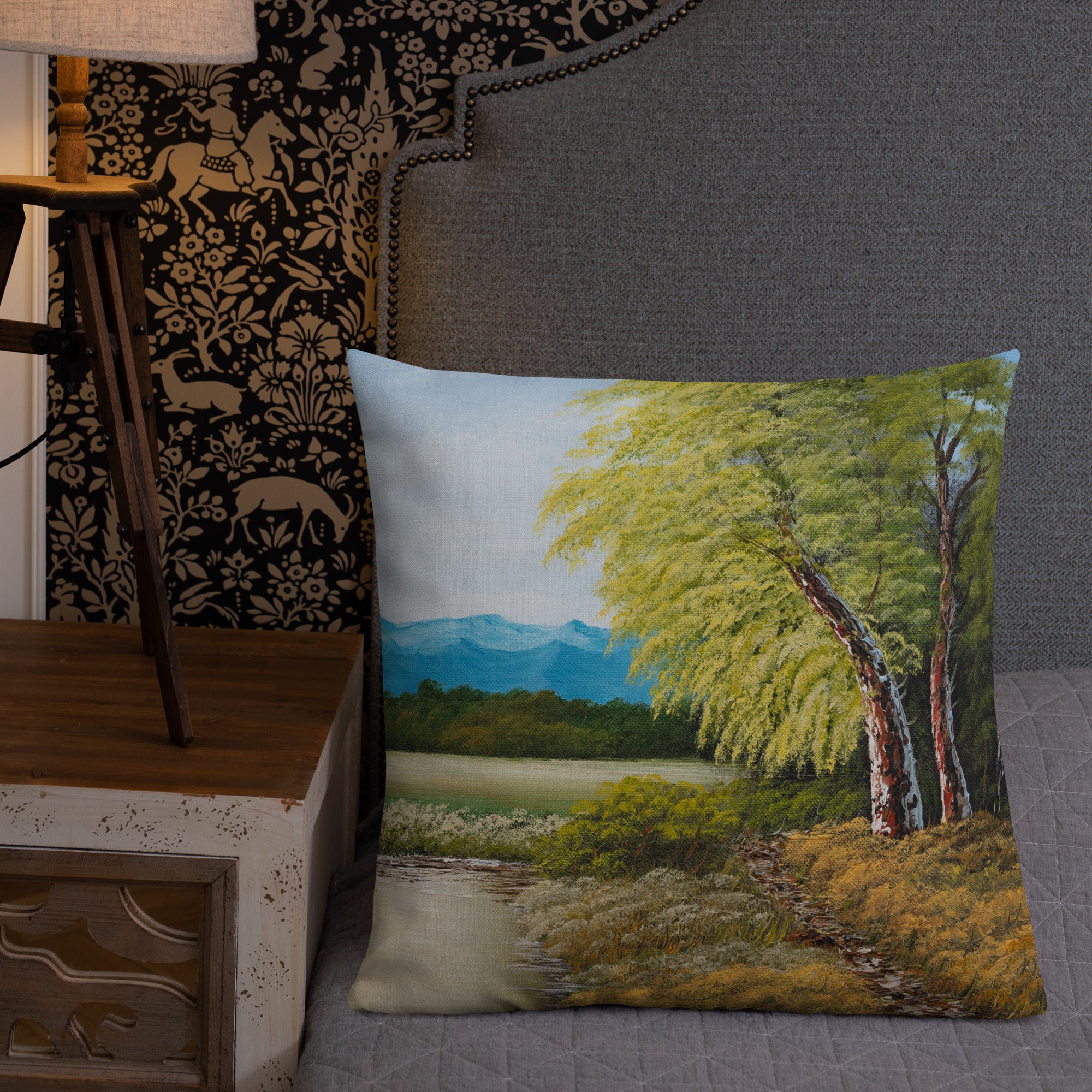 Exclusive and decorative premium designer cushion with an all-over print of a painted landscape with a large glade and a big green tree under a blue sky, placed on a bed.
