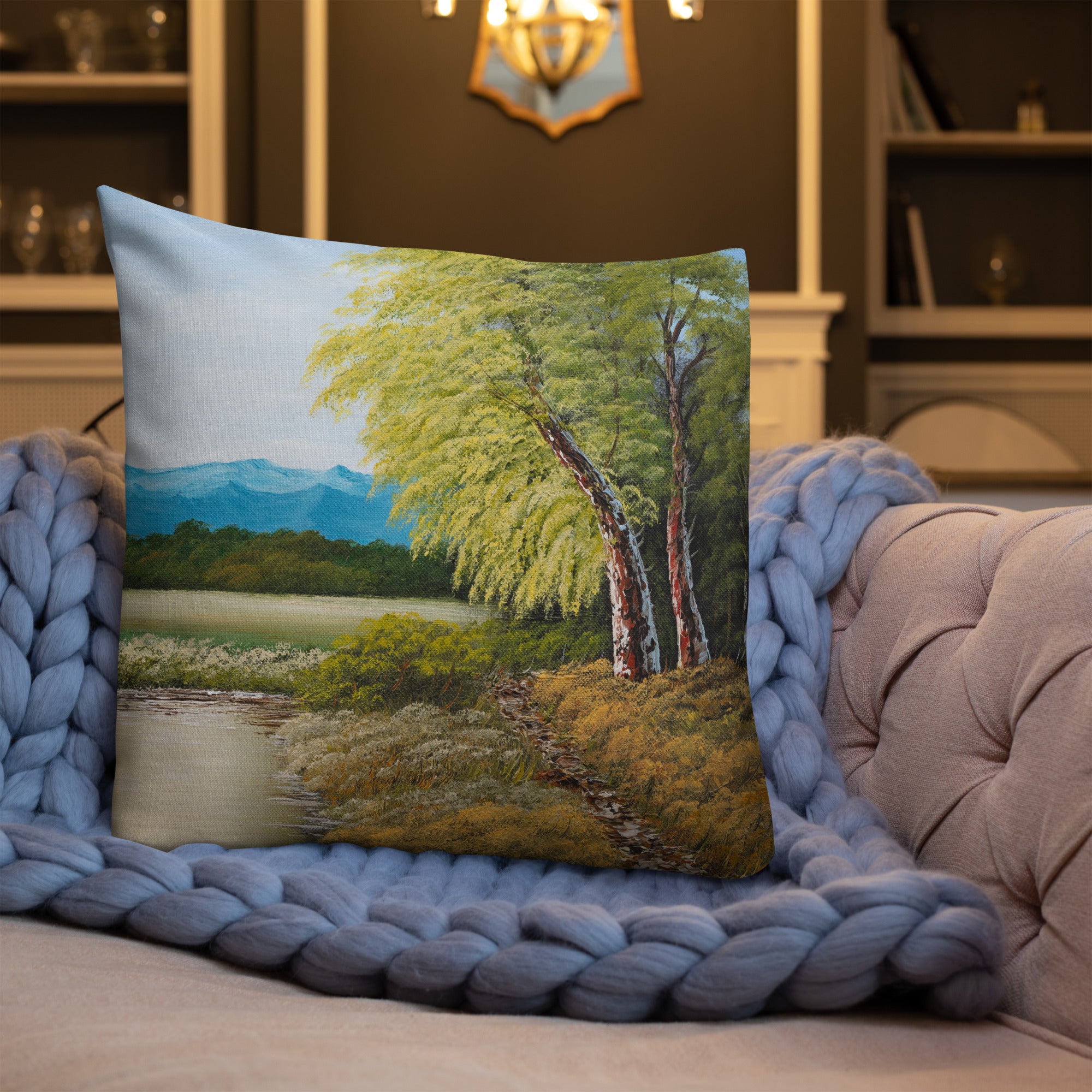 Exclusive and decorative premium designer cushion with an all-over print of a painted landscape with a large glade and a big green tree under a blue sky, placed on a sofa with a purple blanket.