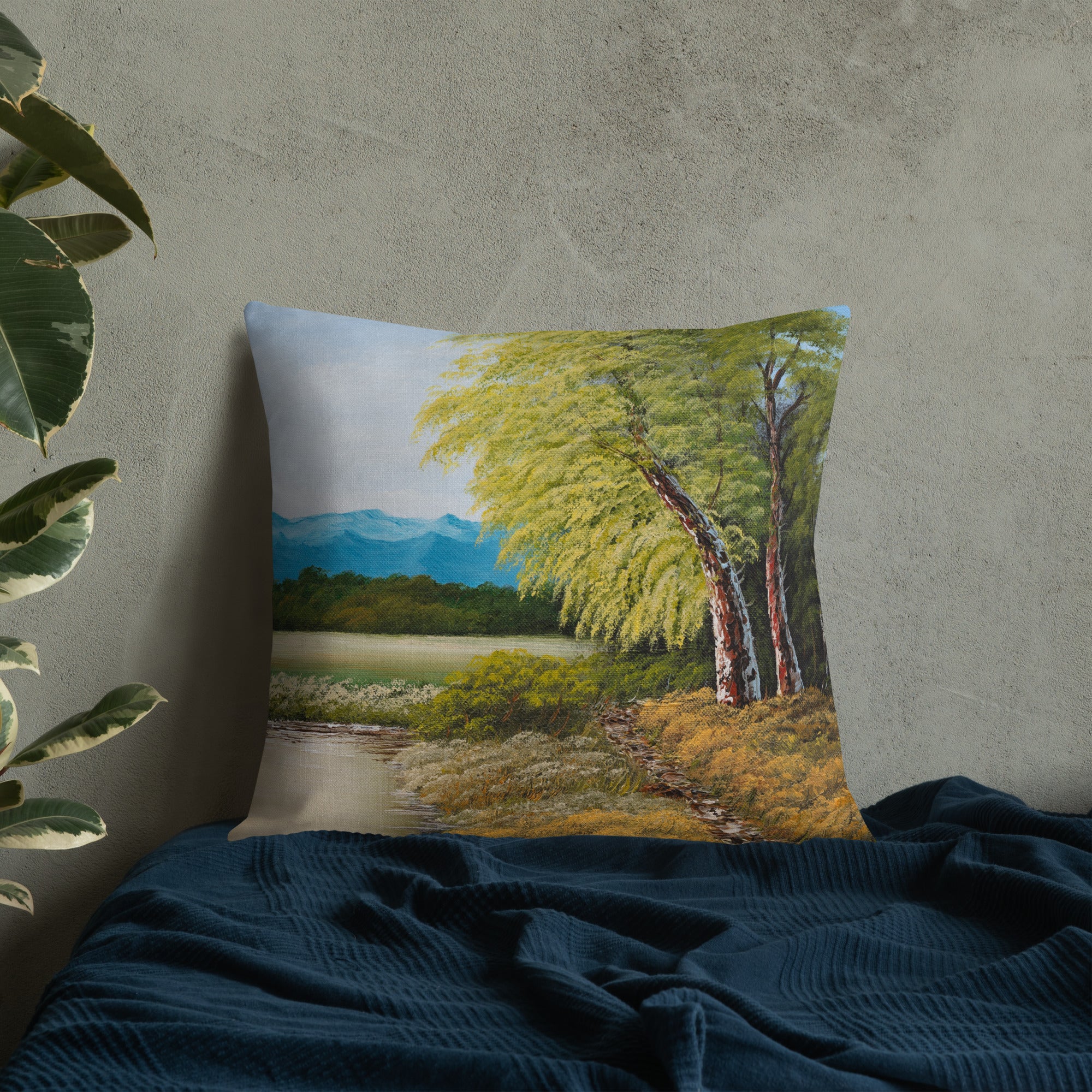 Exclusive and decorative premium designer cushion with an all-over print of a painted landscape with a large glade and a big green tree under a blue sky, placed on a bed.
