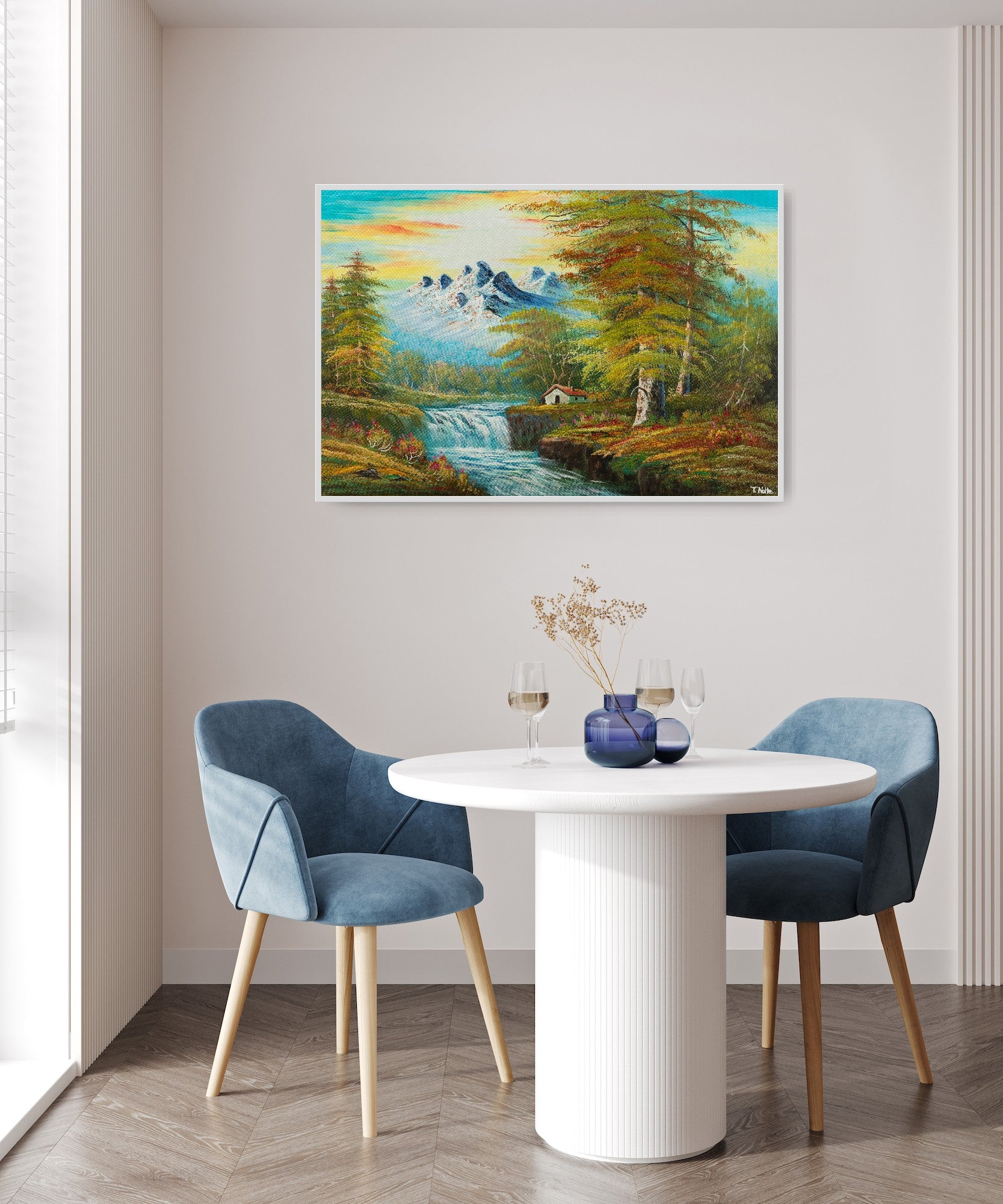 Framed canvas art print of a calm forest landscape with mountains, a cottage next to a river with some waterfalls and lots of trees and bushes, displayed in a modern room with blue chairs.