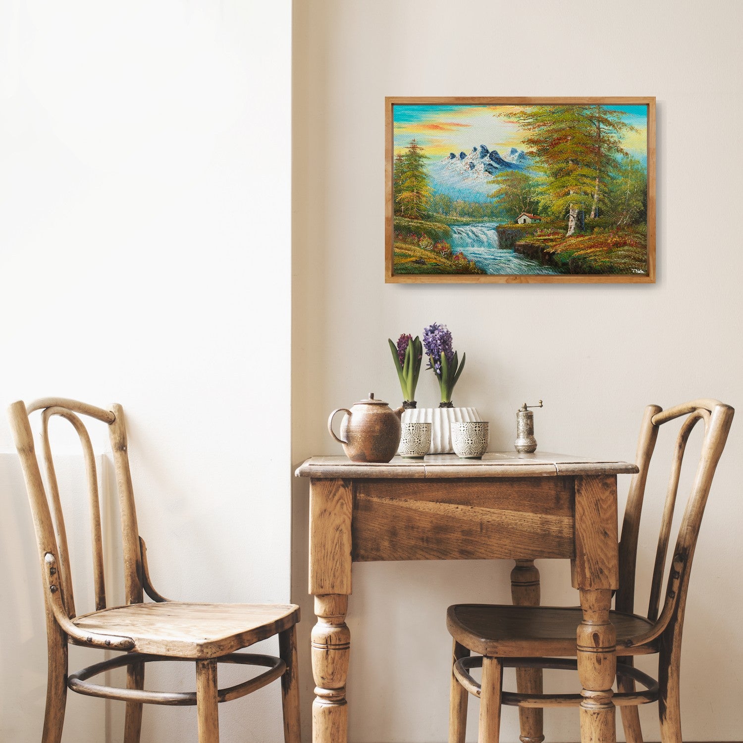 Framed canvas art print of a calm forest landscape with mountains, a cottage next to a river with some waterfalls and lots of trees and bushes, displayed in a kitchen.