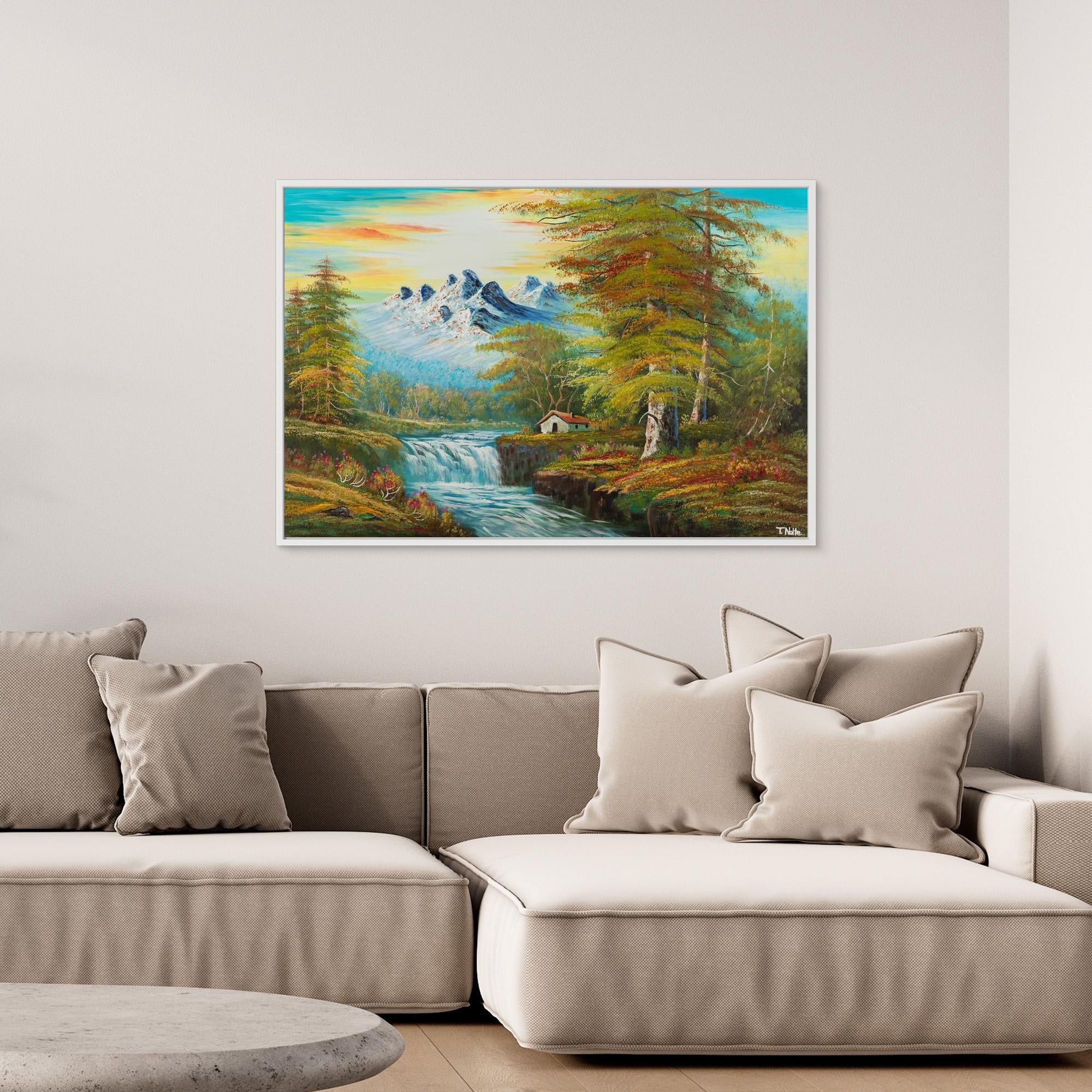 Framed canvas art print of a calm forest landscape with mountains, a cottage next to a river with some waterfalls and lots of trees and bushes, displayed in a modern living room.