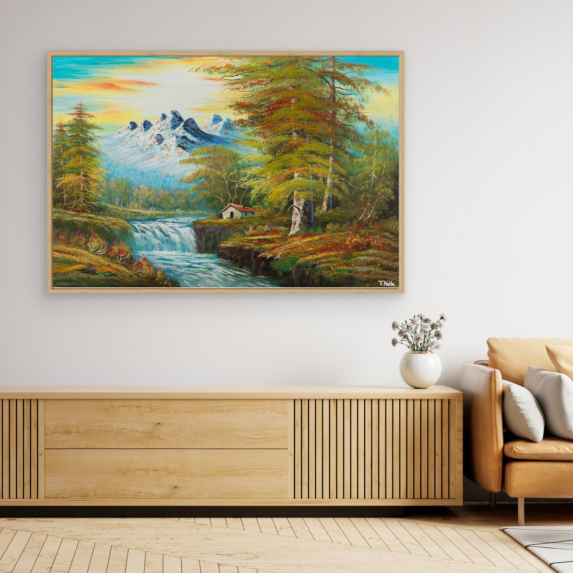 Framed canvas art print of a calm forest landscape with mountains, a cottage next to a river with some waterfalls and lots of trees and bushes, displayed in a modern living room.
