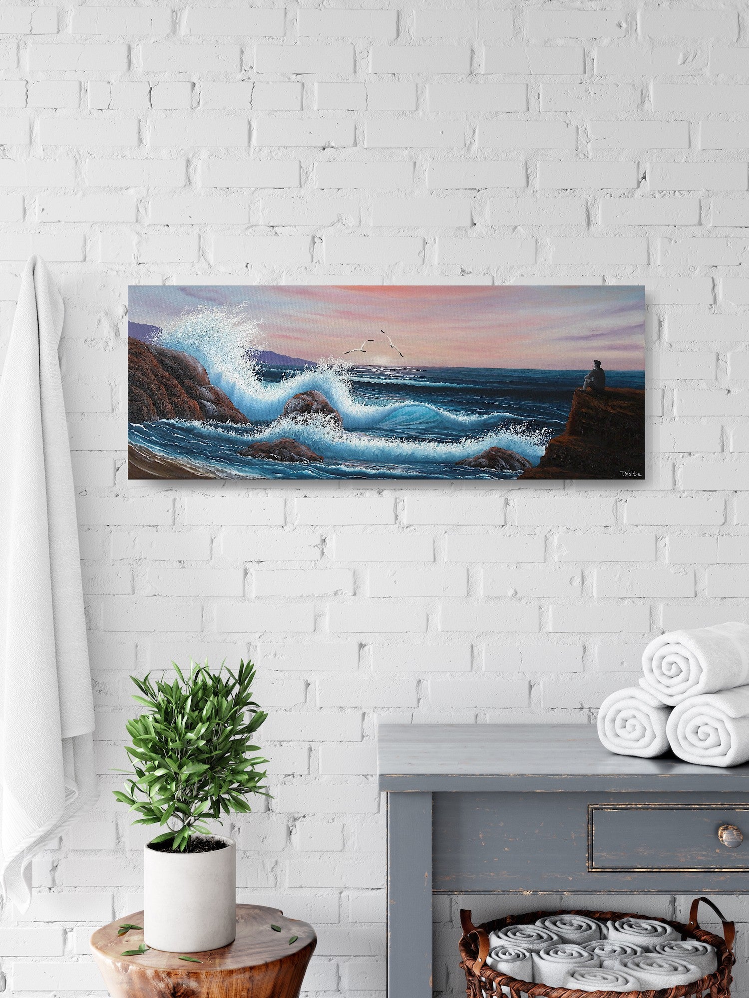 Oil painting on canvas showing a dramatic seascape with large blue waves crashing against rocky cliffs at sunset. Pink and purple clouds fill the sky. A grey person sits on a rock, gazing out at the ocean. The painting is shown in a bathroom.