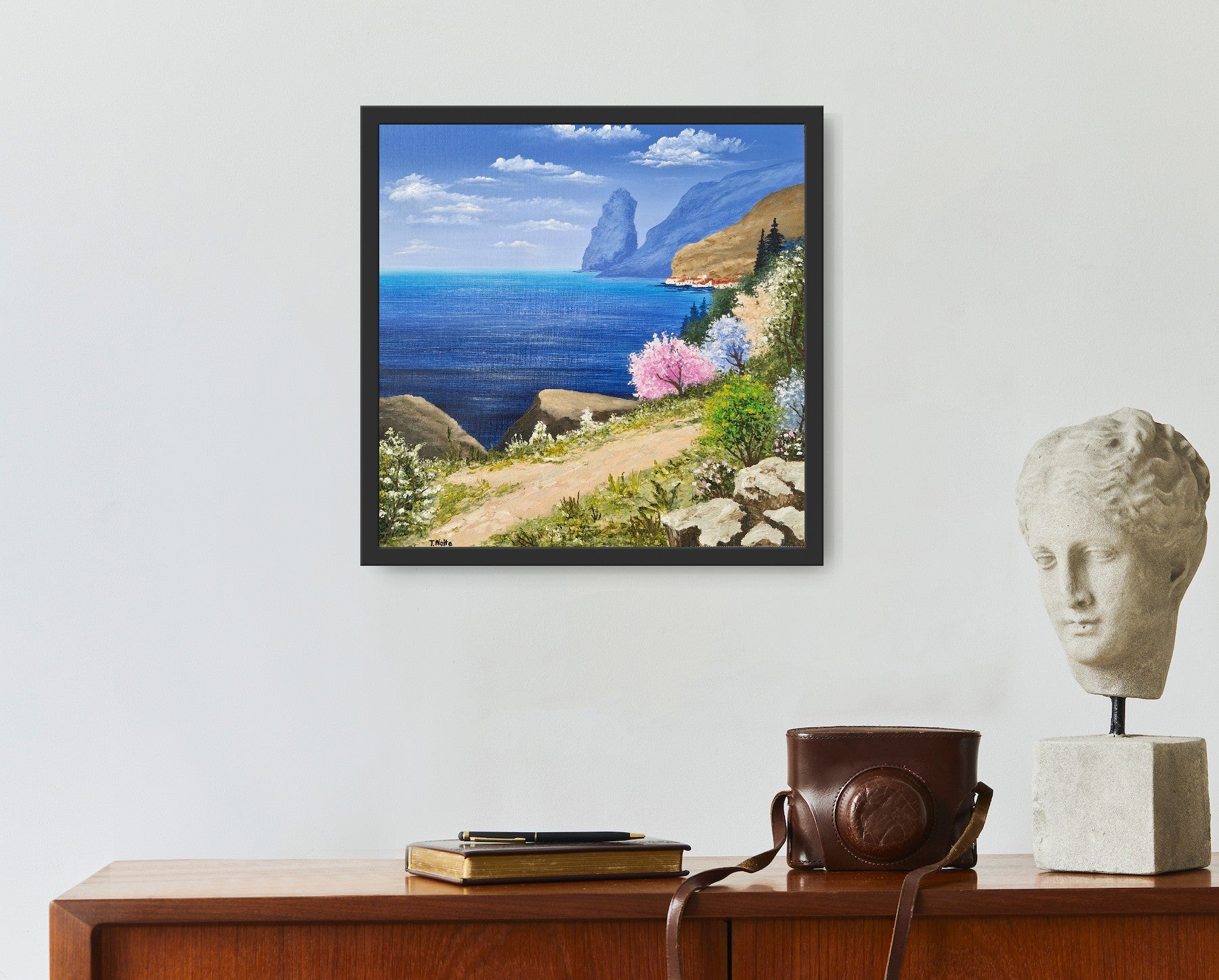 Original artwork by Tobias Nolte titled “Sea View”. A framed, hand-painted oil painting (30x30 cm) featuring a blue sea panorama along a hiking trail, next to colorful trees and rocks, displayed in a modern interior setting.